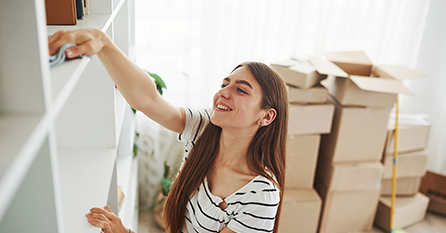 A woman wipes down a shelving unit with moving boxes in the background.