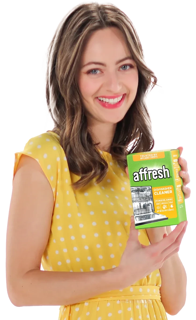A smiling young woman with wavy brown hair, wearing a yellow polka-dot dress, holds up a green and orange box of Affresh Dishwasher Cleaner against a white background.