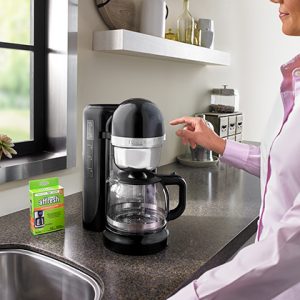 A woman in a pink shirt prepares to press the start button to clean her multi-cup drip coffee machine with an affresh® coffee maker cleaner tablet.