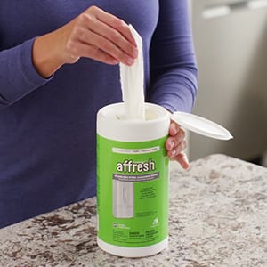 A woman pulls a single affresh Stainless Steel Cleaning Wipe from the top of the 28-count round dispenser so she can clean her stainless steel appliances.