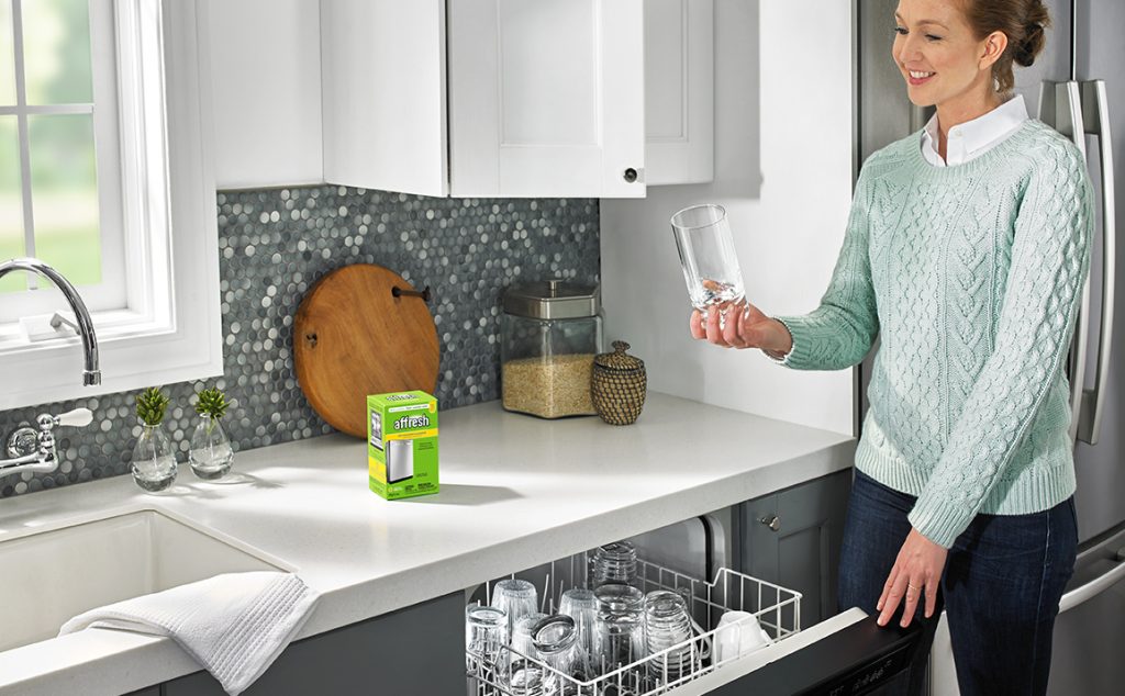 A box of affresh Dishwasher Cleaner tablets sits on the counter while a woman inspects the clean, residue-free glasses she has taken from her dishwasher's top rack.