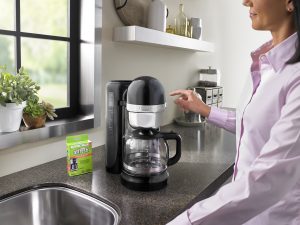 A woman in a pink shirt prepares to press the start button to clean her KitchenAid drip coffee machine with an affresh Coffee Maker Cleaner tablet.