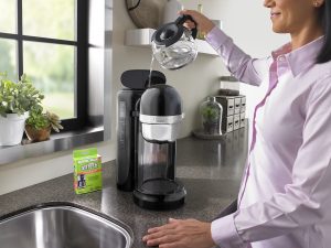 A woman in a pink shirt pours a carafe of water into the reservoir of her KitchenAid coffee machine as she prepares to use an affresh Coffee Maker Cleaner tablet.