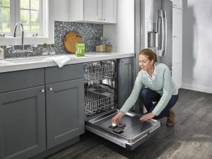 A woman kneels on the floor in her kitchen, placing an affresh® dishwasher cleaner tablet into her dishwasher's detergent dispenser while a box of affresh sits nearby.