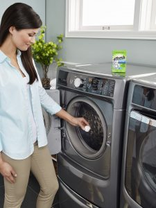 A long-haired woman in an aqua shirt places an affresh® washing machine cleaner tablet into the drum of her front-load washer and prepares to run the "Clean with affresh" cycle. A box of affresh® washing machine cleaner sits atop the washer.