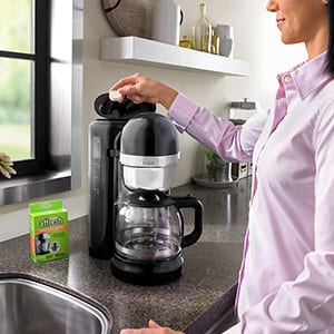A woman in a pink shirt drops a single affresh Coffee Maker Cleaner tablet into the reservoir of her drip coffee machine to remove mineral deposits and other residue.