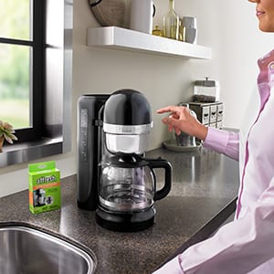 A woman in a pink shirt prepares to press the start button to clean her drip coffee machine with an affresh Coffee Maker Cleaner tablet.