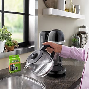 A woman in a pink shirt pours the carafe of affresh Coffee Maker Cleaner solution into the sink after using the tablet to clean her KitchenAid coffee machine.