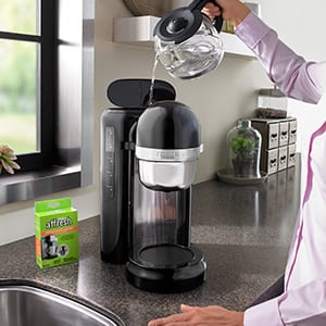 A woman in a pink shirt pours the carafe of affresh Coffee Maker Cleaner solution into the sink after using the tablet to clean her KitchenAid coffee machine.