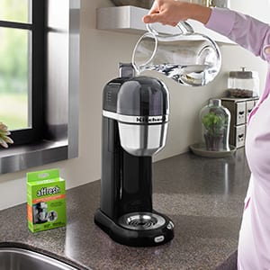 A woman in a pink shirt pours a glass pitcher of water into the reservoir of a KitchenAid personal coffee machine as she prepares to clean it with an affresh Coffee Maker Cleaner tablet.