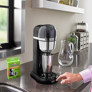 A woman prepares to press the start button on her KitchenAid personal coffee machine to clean and descale it with an affresh Coffee Maker Cleaner tablet.