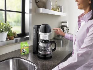 A woman in a pink shirt prepares to press the start button to clean her KitchenAid drip coffee machine with an affresh Coffee Maker Cleaner tablet.