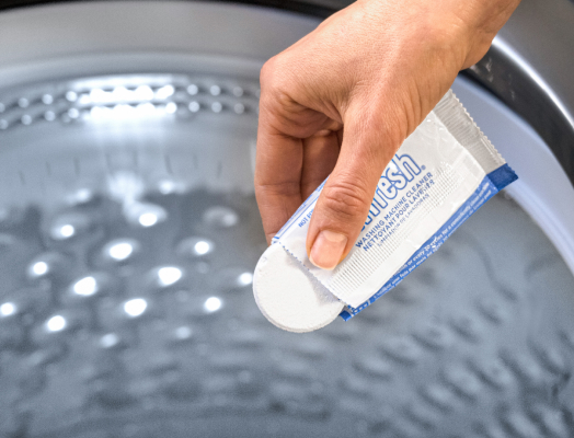 A person dropping an affresh® washing machine cleaner tablet into a washing machine drum for deep cleaning.