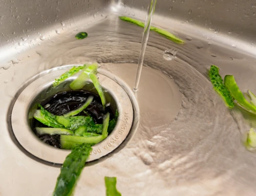 Close-up of green vegetable scraps, including celery and cucumber peels, clogging a stainless steel sink drain labeled "Food Waste Disposer," with water streaming down from above.