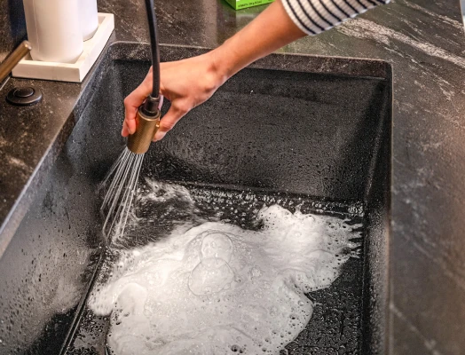 A hand in a striped sleeve holding a black pull-out kitchen faucet spraying water into a dark stone sink, creating white foam and bubbles at the bottom.