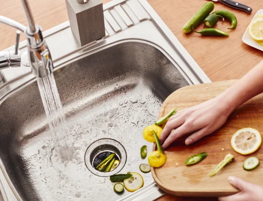 Overhead view of a person's hand scraping chopped cucumbers, lemons, and peppers from a wooden cutting board into a stainless steel kitchen sink with running water from a chrome faucet, surrounded by scattered vegetable pieces.