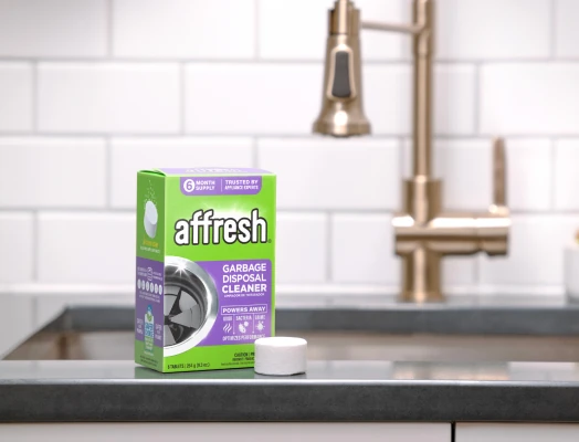 A green box of affresh Garbage Disposal Cleaner tablets placed on a dark kitchen countertop, with a single round white tablet in front, and a brushed gold faucet and white tiled backsplash in the background.
