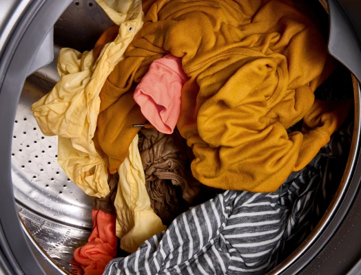 Overhead view inside a washing machine drum filled with a tangled pile of colorful clothing items in yellows, oranges, browns, pinks, and striped patterns.