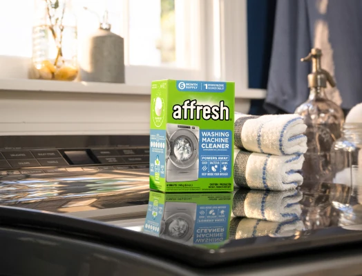 A box of affresh Washing Machine Cleaner tablets placed on a dark countertop in a laundry area, with a single blue tablet in front, folded blue-striped towels beside it, and background elements like hanging clothes, a soap dispenser, and potted plants.