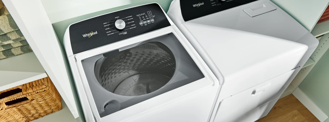 Overhead view of a white Whirlpool top-load washing machine with its lid open, revealing a perforated drum, positioned next to a matching white dryer in a laundry closet with light green walls, wooden shelves holding folded towels, and a wicker basket below.
