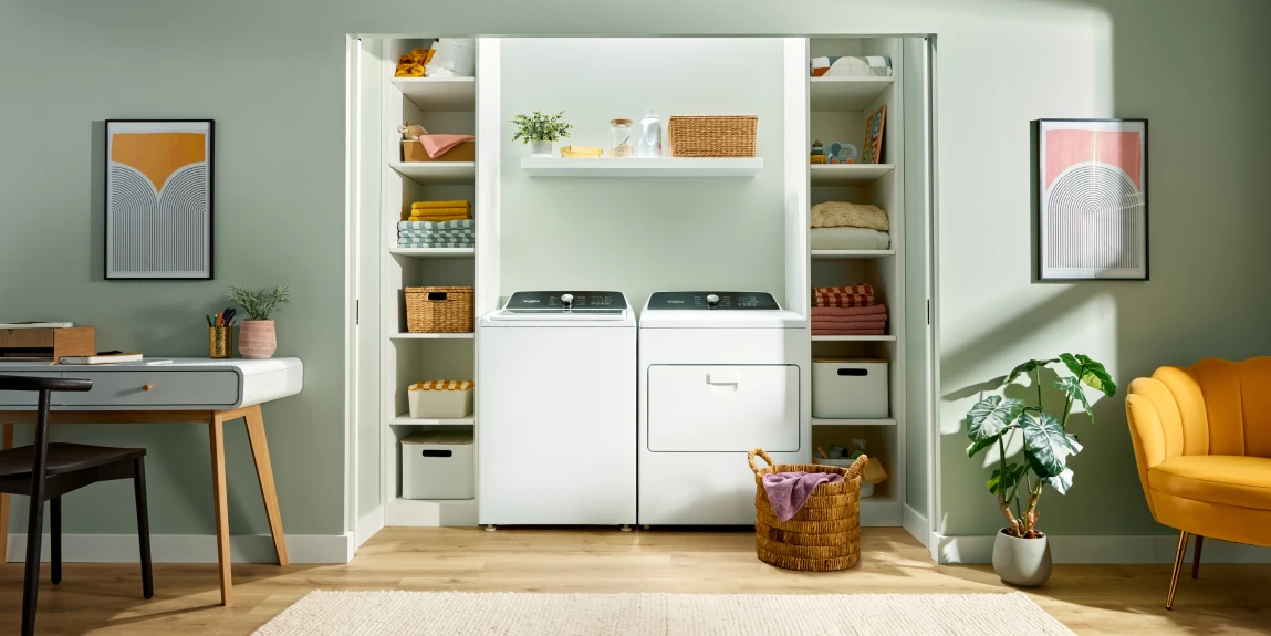 A neatly organized laundry closet in a light green room featuring a white top-load washer and front-load dryer side by side, flanked by shelves with baskets, folded linens, and decor; a wooden desk with chair on the left, potted plants, a yellow armchair on the right, and abstract wall art.