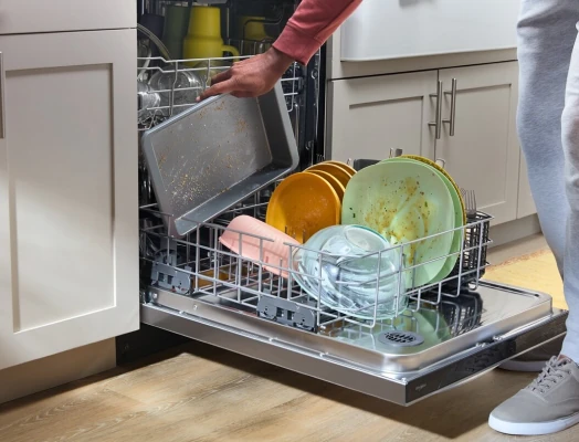 A person in a red sleeve and gray pants loads a greasy baking sheet into the lower rack of an open dishwasher filled with dirty plates, bowls, glasses, and utensils covered in food residue.