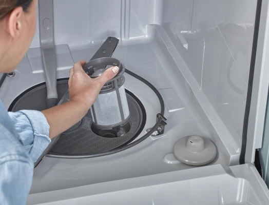 A close-up of a woman's hand in a blue shirt holding a removable cylindrical mesh filter inside the bottom of an open dishwasher, with the spray arm, drain area, and other interior components visible.