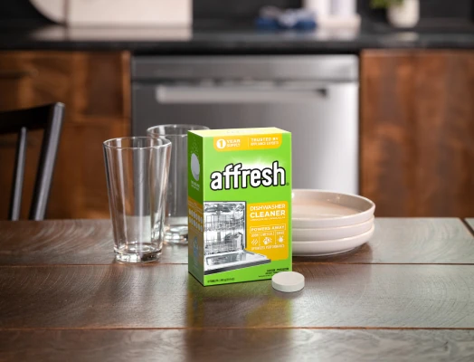 A box of affresh Dishwasher Cleaner tablets sits on a wooden table next to a single round white tablet, two clear drinking glasses, and a stack of white plates, with a stainless steel dishwasher visible in the background of a kitchen setting.