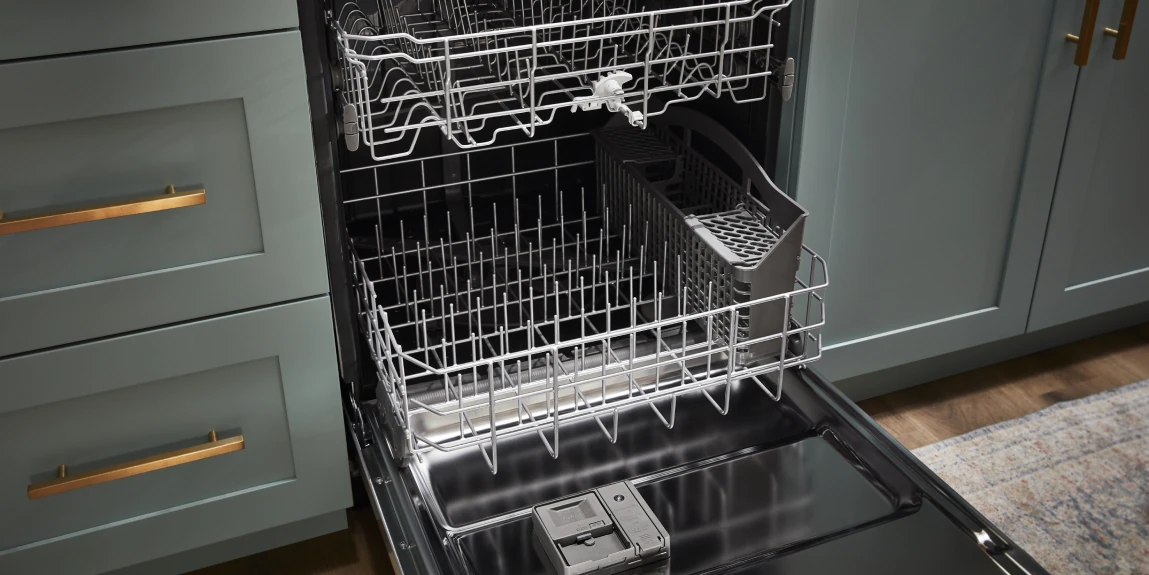 Overhead view of an empty open dishwasher with the upper and lower wire racks extended, including a removable silverware basket and detergent dispenser, set in a kitchen with teal cabinets and brass handles.