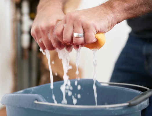 A man's hands, one wearing a silver ring, squeeze a yellow sponge, causing soapy water to drip into a blue plastic bucket below, with plumbing pipes visible in the background.
