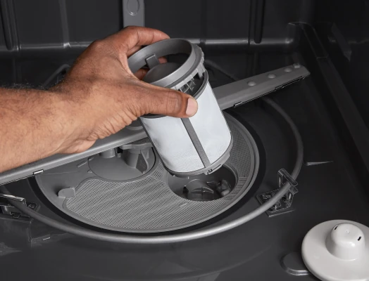 A hand holds a removable cylindrical mesh filter extracted from the bottom of an open dishwasher, with the interior spray arm, drain, and other components visible.