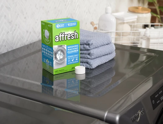 A blue box of affresh Washing Machine Cleaner tablets placed on top of a gray washing machine, with a single round white tablet in front and a stack of folded blue towels beside it, in a laundry room setting with baskets and bottles in the background.