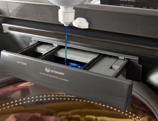 Close-up of blue liquid detergent being poured from a white bottle into the "Main Wash" compartment of a pulled-out gray dispenser drawer on a washing machine, labeled for softener and detergent.