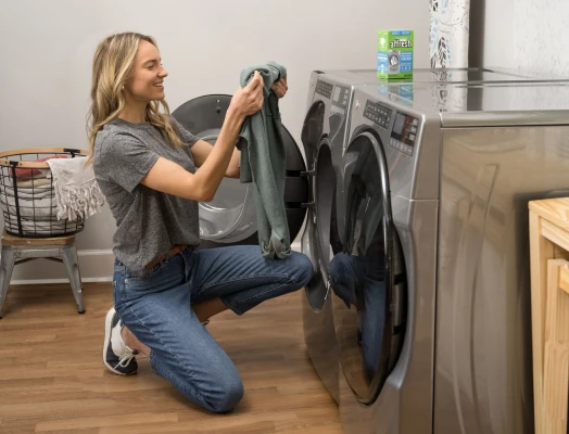 A smiling woman in a gray shirt and jeans kneels to remove a green garment from an open front-load stainless steel washing machine, with a matching dryer beside it, an affresh cleaner box on top, and laundry baskets nearby in a wooden-floored room.