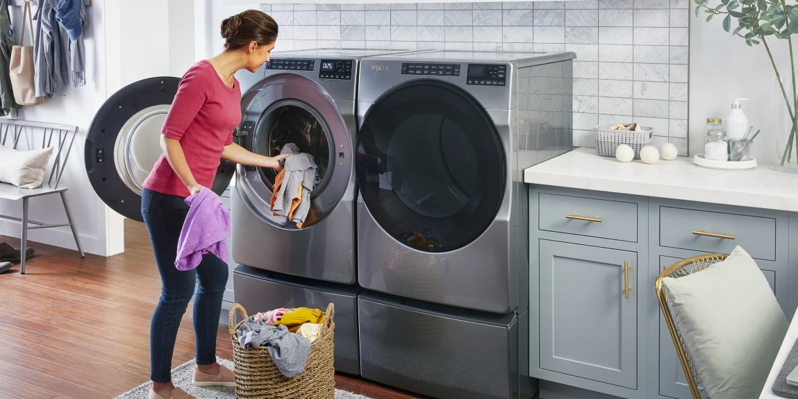 A woman in a pink shirt and jeans transfers wet clothes from an open front-load washer to a dryer in a modern laundry room with gray appliances, a wicker basket of laundry at her feet, hanging clothes, marble tiled backsplash, and decor like plants and wool dryer balls on the counter.