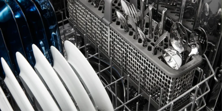 Close-up of a dishwasher rack with white plates, a blue pan, and a silverware basket of utensils